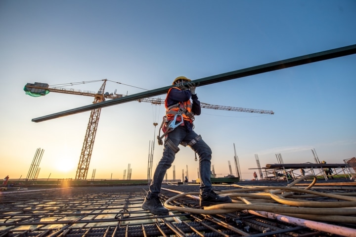 construction worker carrying long plank of wood across site